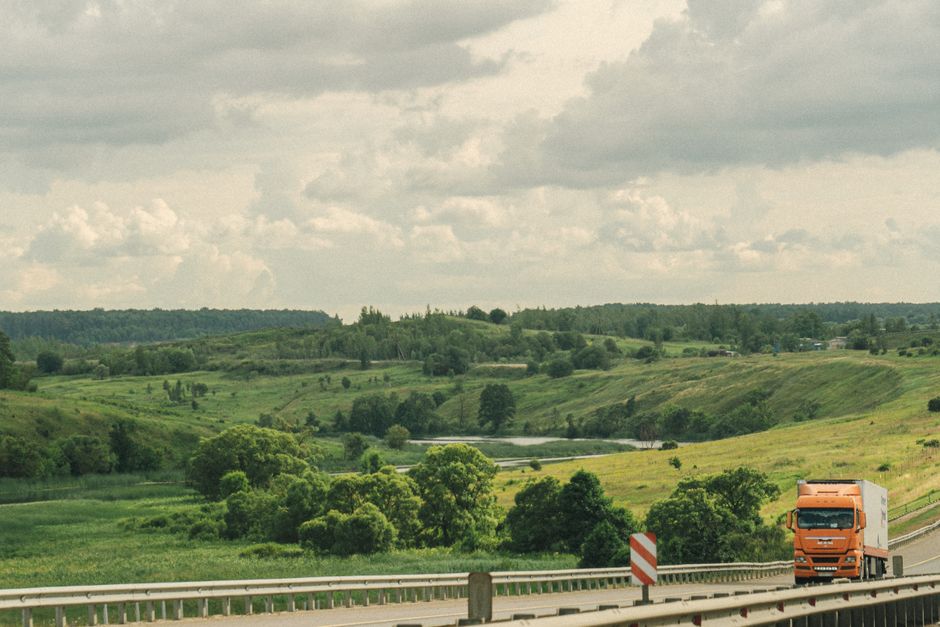 An orange truck drives along a highway through a lush, green, hilly countryside under a cloudy sky.